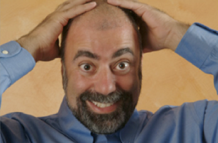 Daniel Klein as he is today - shaved head, trimmed beard, hands on head, wearing blue shirt, smiling