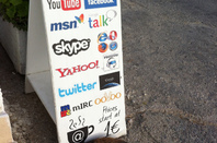 Cat lies exhausted in front of a sandwich board advertising an internet cafe, with all manner of tech companies' branding plastered all over it: Google, Yahoo, Facebook, MSN , Skype, ,Opera, Ventrilo, Twitter, MIRC!!!