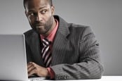 Man looks suspiciously over his shoulder while working on laptop. Photo via Shutterstock