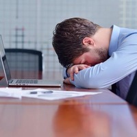 Defeated-looking young man puts his head against table in front of laptop and pile of papers in conference room. Pic via Shutterstock
