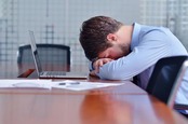 Defeated-looking young man puts his head against table in front of laptop and pile of papers in conference room. Pic via Shutterstock