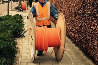 Man rools out fibre cable from a large wooden cable reel on a suburban street. Pic via Pixabay