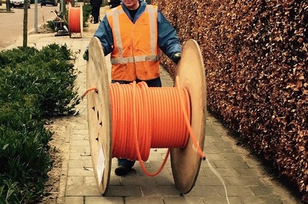Man rools out fibre cable from a large wooden cable reel on a suburban street. Pic via Pixabay