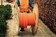 Man rools out fibre cable from a large wooden cable reel on a suburban street. Pic via Pixabay