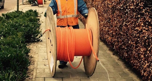 Man rools out fibre cable from a large wooden cable reel on a suburban street. Pic via Pixabay