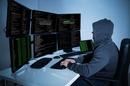 Hacker with face obscured, wearing a hoodie,  works in front of a bank of monitors. photo by Shutterstock