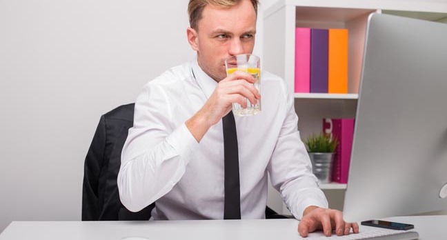 Man types something into Mac while sipping a glass of lemon water. Not a brilliant idea. Photo by SHutterstock