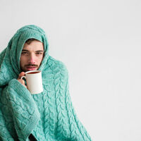 Man wrapped in turqouise cable-knit blanket sips from hot drink - is clearly ill. Photo by Shutterstock