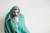 Man wrapped in turqouise cable-knit blanket sips from hot drink - is clearly ill. Photo by Shutterstock