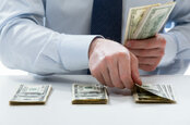 Man in suit counts out piles of dollars. Photo via Shutterstock