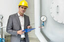Man with clipboard, hardhat and concerned expression next to a pressure gauge in an industrial setting. Pic by Shutterstock