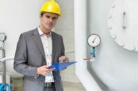 Man with clipboard, hardhat and concerned expression next to a pressure gauge in an industrial setting. Pic by Shutterstock