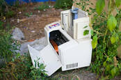 ewaste: a broken printer lies abandoned in an alleyway, weeds growing through it.