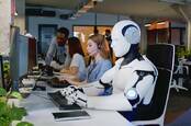 A humanoid robot working at a PC at a desk next to humans