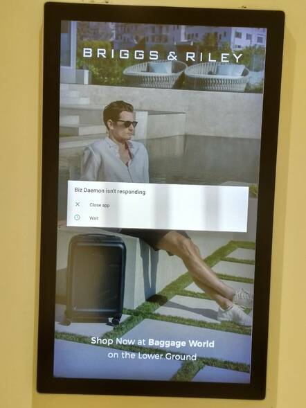 Electronic signage with pop-up text "Biz Daemon is not responding" Above an image of a man in shorts sitting against a wall with a wheeled suitcase